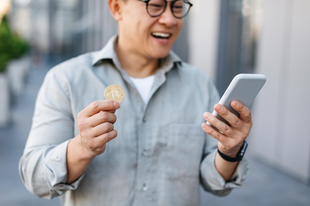 Online trading Happy asian man holding golden bitcoin and smartphone middle aged male earning cryptocurrency