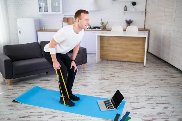 Online training. Young man doing exercise with fitness rubber bands with online tutorial at home, free space. Doing sports at home