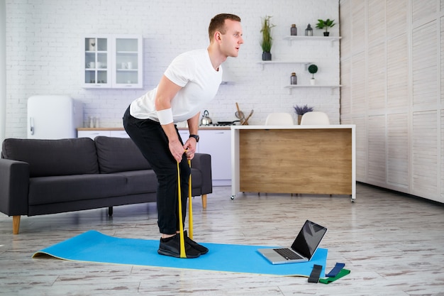 Online training. Young man doing exercise with fitness rubber bands with online tutorial at home, free space. Doing sports at home