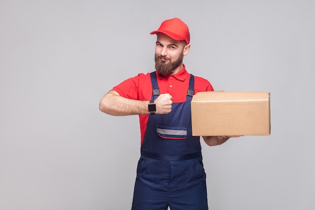 Ontime delivery service! Young logistic bearded man in blue uniform and red t-shirt standing, holding cardboard box and showing watch on grey background. Indoor, studio shot, isolated, copy space