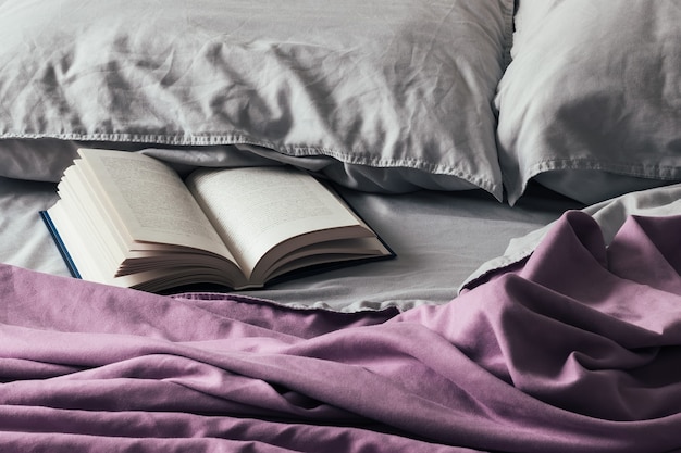 Open book on bed with purple bedspread and gray pillows. Fascinating book in soft crumpled bedding