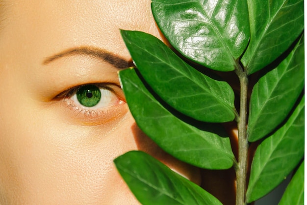 An open green eye of a woman and a plant with large green leaves covering the second eye