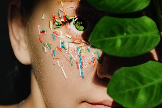 An open green woman's eye with a sweet multicolored sprinkle on the eyelid and a plant with large green leaves