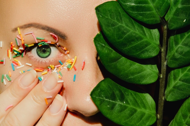 An open green woman's eye with a sweet multicolored sprinkle on the eyelid and a plant with large green leaves