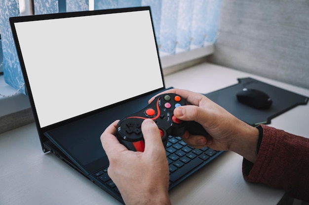 Open laptop computer with a blank white screen with copy space standing open on table alongside a video game controller indoors at home joystick for playing in men's hands