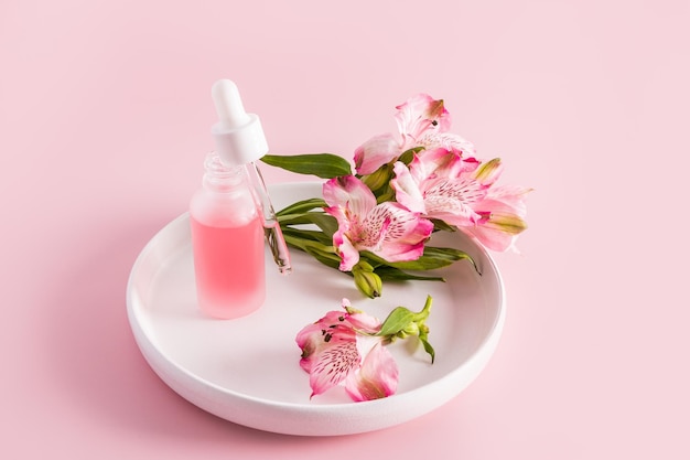 An open matte white bottle with a filled pipette of natural moisturizer facial skin care stands on a white ceramic plate pink background