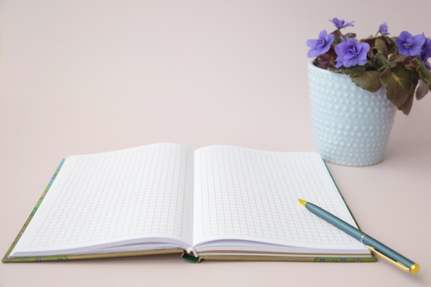 An open notebook in a cage with a pen for writing next to a violet on a pink background a place for text template