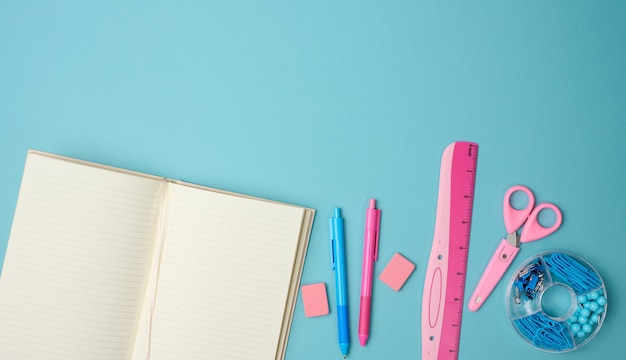 Open notebook with blank pink sheets, scissors and ruler on blue background, top view