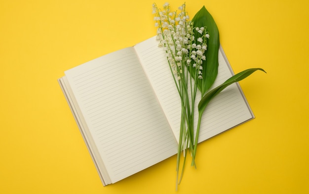 Open notebook with blank white sheets and a bouquet of blooming lilies of the valley on a yellow surface, top view