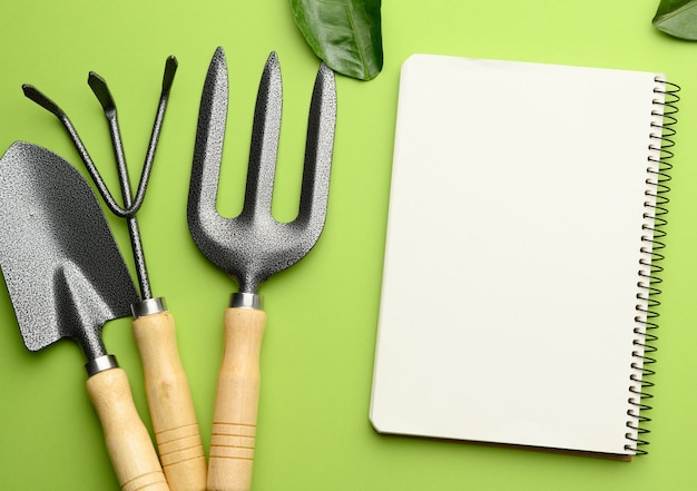 Open notebook with blank white sheets and various gardening tools with wooden handles on green background, flat lay, copy space