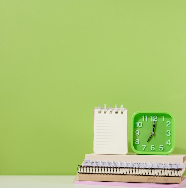 An open notepad and a green alarm clock stands on a stack of notebooks a green background