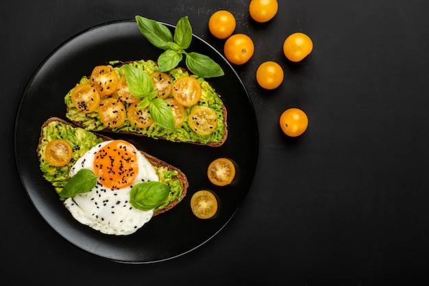 Photo open sandwiches with avocado guacamole, yellow cherry tomatoes, fried egg and basil on a black plate . top view. copy space.