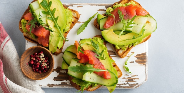 Open toast with trout, salmon, avocado, cucumber and arugula on wooden stand on gray concrete background. Selective focus. Top view. Copy space