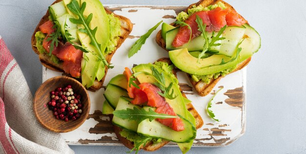 Photo open toast with trout, salmon, avocado, cucumber and arugula on wooden stand on gray concrete background. selective focus. top view. copy space