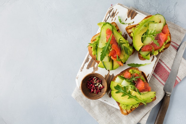 Open toast with trout, salmon, avocado, cucumber and arugula on wooden stand on gray concrete background. Selective focus. Top view. Copy space