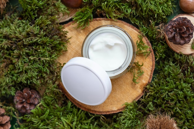 Opened cream jar with white cap on wooden piece between green moss top view mockup