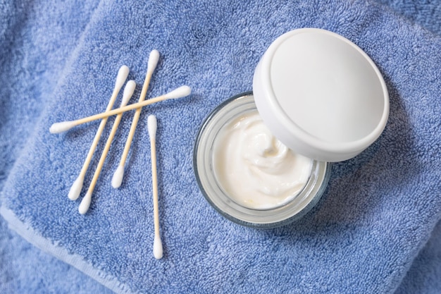 Opened cream jar with white lid near cotton swabs on blue towel top view cosmetic mockup