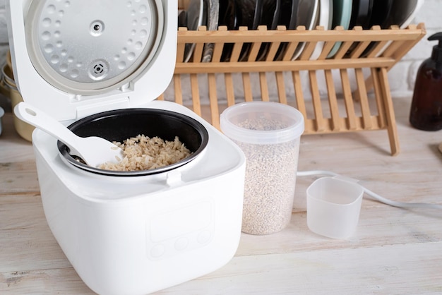 Opened electric rice cooker with cooked steaming brown rice on wooden countertop in the kitchen