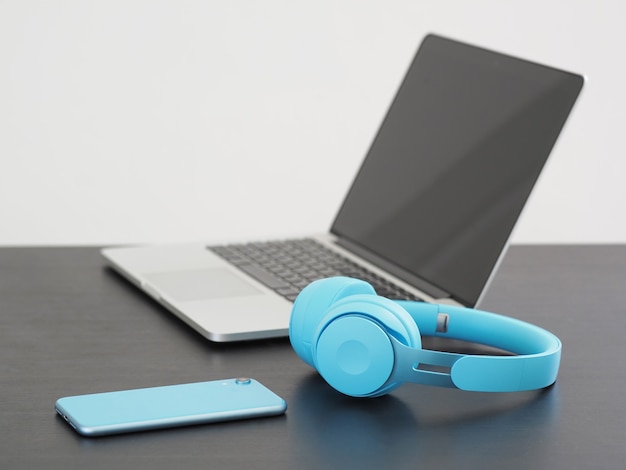 Opened laptop with smartphone and blue wireless headphones on wooden desk. Selective focus.