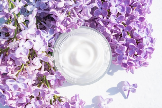 Opened white jar of herbal light cream for women on white table Beautiful branch of purple lilac Care for clean and soft face hands legs and body skin Fresh flowers Empty place for text