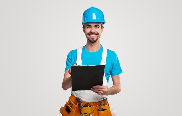 Optimistic adult bearded male contractor in hardhat with toolkit and clipboard smiling and looking at camera against white background