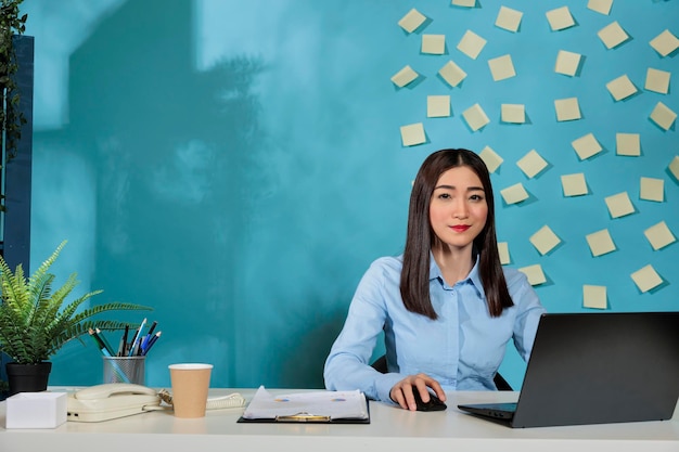 Optimistic asian woman working from a modern office with laptop with reminder sheets taped to the wall. Businesswoman works with portable computer from company workplace.