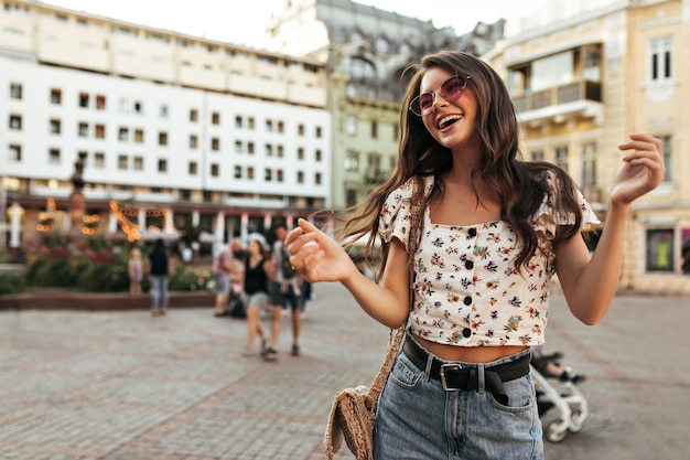 Optimistic brunette woman in jeans and summer stylish blouse dance outdoors Curly young girl in denim pants floral top and pink sunglasses smile and move on street