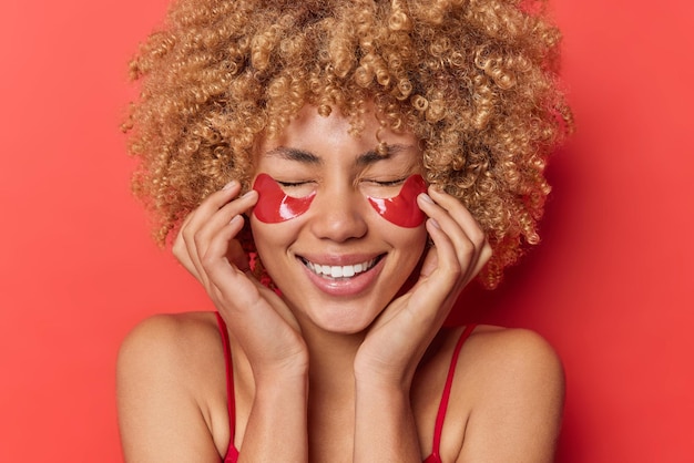 Optimistic cheerful curly haired woman applies collagen patches under eyes to reduce dark circles and wrinkles undergoes beauty procedures poses against vivid red background. Skin care concept