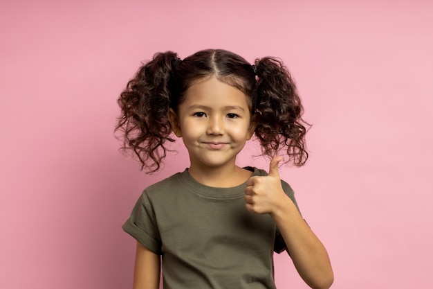 Optimistic cheerful small girl with curly funny hairstyle, showing thumb up, like gesture, expressing approval, wearing casual t shirt, smiling, standing. Body language.