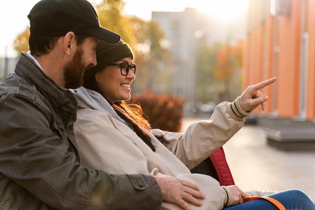 Optimistic man embracing his lovely woman while she pointing down the street with interest during the sitting at the bench at the street