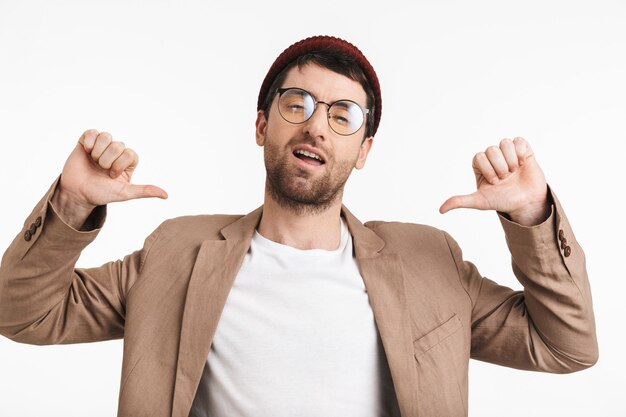 Photo optimistic man with stubble wearing hat and eyeglasses pointing thumbs at himself isolated over white wall