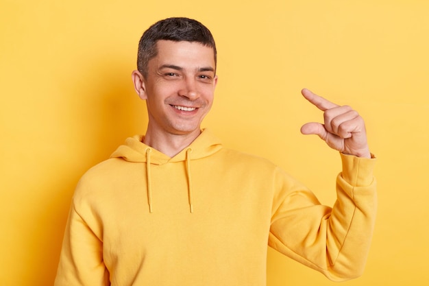 Optimistic positive man wearing casual style hoodie posing isolated over yellow background showing a little bit gesture inch or centimeter disappointed with minimum size