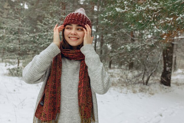 Optimistic woman adjusting warm hat while standing in snowy coniferous forest in winter