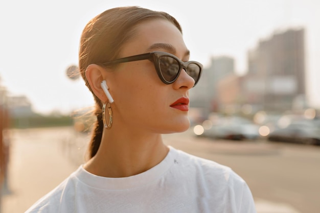 Optimistic woman with dark hair in white tshirt listening music in city Cool lady in light blouse and black sunglasses looking aside on city