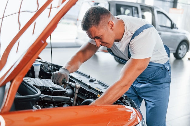 Orange colored car Man in uniform is repairing broken automobile indoors