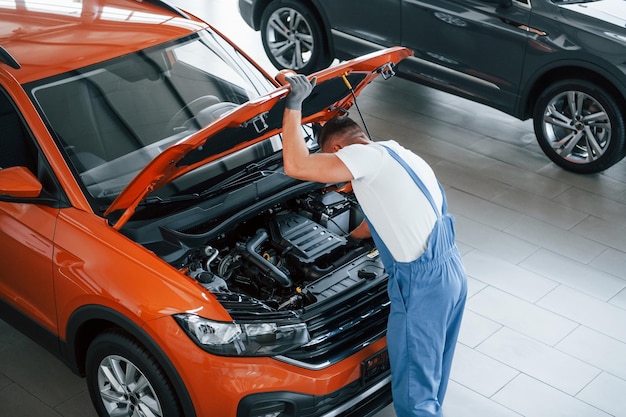 Orange colored car Man in uniform is repairing broken automobile indoors