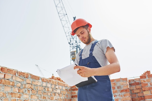 In orange colored hard hat Young construction worker in uniform is busy at the unfinished building