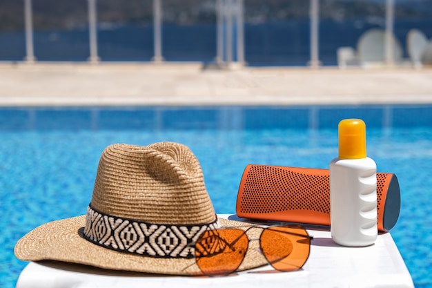 Orange coloured beach accessories near swimming pool. Sun cream, sunglasses, music speaker and straw hat.