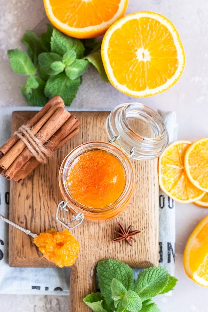 Orange jam in glass jar, orange fruit slices, mint and spices on light grey