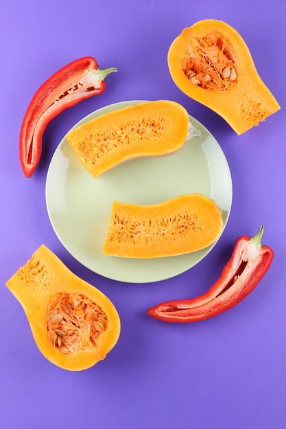 Orange juicy pumpkin halves with red pepper halves on a white plate Closeup of colorful vegetables on a purple background View from above