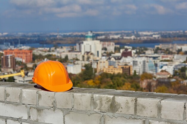 Orange plastic safety helmet is on a brick wall roof of modern building under construction blurred panoramic cityscape blue sky sunny day