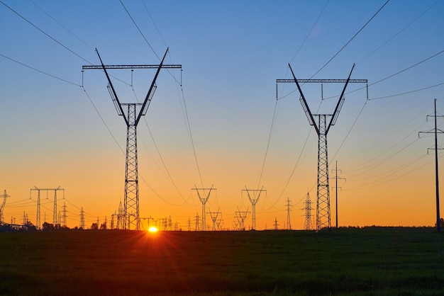 Orange sunrise on field with high voltage electricity towers Dark silhouettes of repeating power lines on sunrise Electricity generation transmission and distribution network Industrial landscape