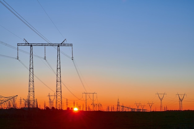 Orange sunrise on field with high voltage electricity towers Dark silhouettes of repeating power lines on sunrise Electricity generation transmission and distribution network Industrial landscape