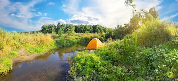 Orange tourist tent on the bank of the river with the banks covered with greenery. Sunny day