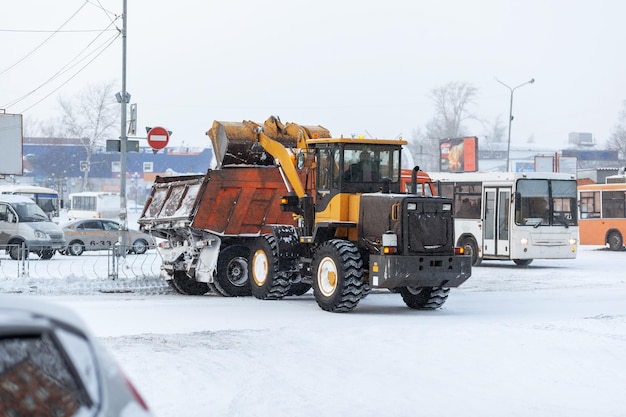 Orange tractor cleans up snow from the road and loads it into the truck. Cleaning of roads