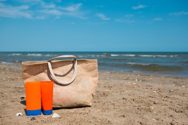 An orange tube with mock up sunscreen sunglasses and a beach bag stand on the sand near the sea and swing from the breeze UV protection beach holidays travel