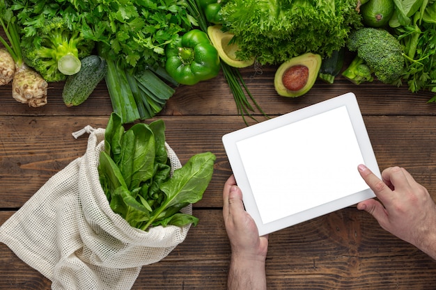 Order food online Man holds tablet with blank screen for your text message or design on wooden table with fresh green vegetables and textile bag