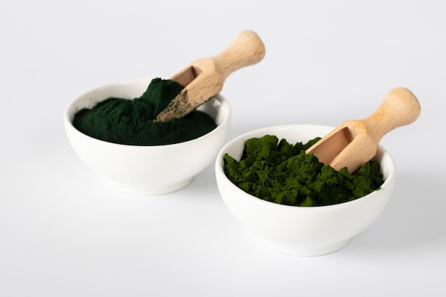 Organic algae chlorella and spirulina in powder form in white bowls with wooden scoops on white background.