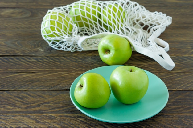 Organic Granny Smith apples on a plate with mesh textile bag on wooden table.