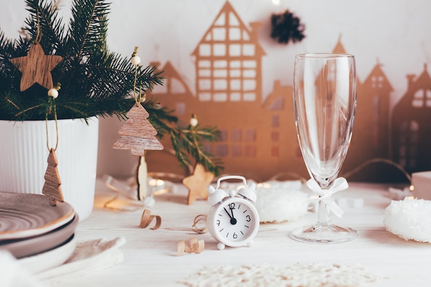 Organization of a festive Christmas table. White alarm clock and a glass of champagne on the background of a fir bouquet in a vase and a cardboard house. Tablescapes on white wooden table